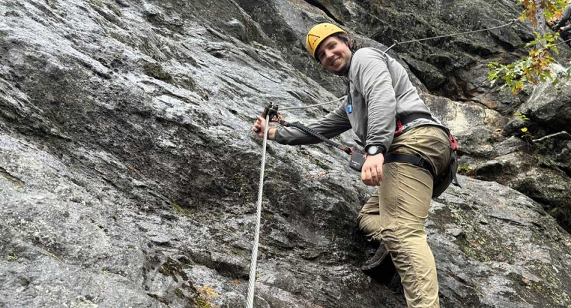 A person wearing safety gear is secured by ropes as they look down at the camera while rock climbing